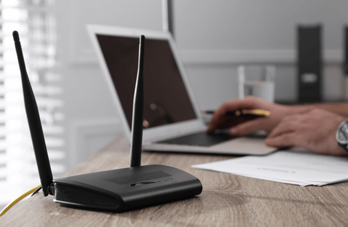 A wireless router (CPE) on top of a desk next to hand typing on a laptop computer.